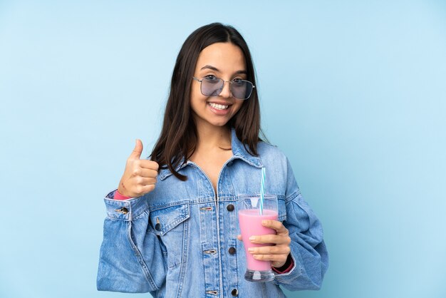 Jonge vrouw met aardbeimilkshake over blauw die ok teken en duim op gebaar tonen