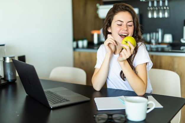 Jonge vrouw met appel en laptop zitten in de keuken