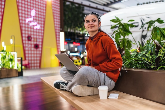 Jonge vrouw met behulp van haar laptop zittend op een bank glimlachend en wegkijkend in een winkelcentrum