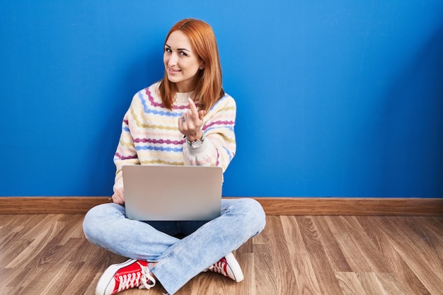 Jonge vrouw met behulp van laptop thuis zittend op de vloer wenkend kom hier gebaar met de hand uitnodigend verwelkomen blij en glimlachend