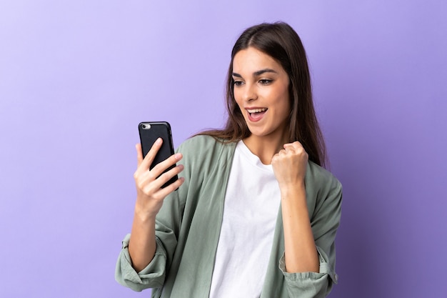 Jonge vrouw met behulp van mobiele telefoon geïsoleerd op paarse muur een overwinning vieren