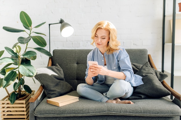 Jonge vrouw met behulp van mobiele telefoon op gezellige zwarte bank, woonkamer in witte tinten en pot met bloem