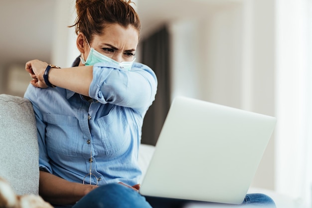 Jonge vrouw met beschermend masker die in de elleboog niest tijdens het gebruik van de computer thuis