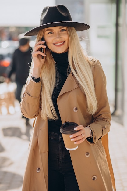 Jonge vrouw met blond haar die zwarte hoed draagt die aan de telefoon spreekt en koffie drinkt