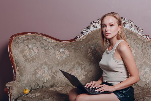 Jonge vrouw met blond haar zit en gebruikt laptop op de bank