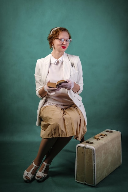 Foto jonge vrouw met bril gekleed in retro stijl zittend op een stoel met koffer aan haar voeten