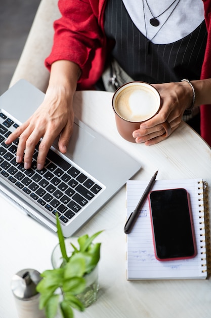 Jonge vrouw met cappuccino toetsen van laptop toetsenbord in te drukken tijdens het netwerken door tafel in café