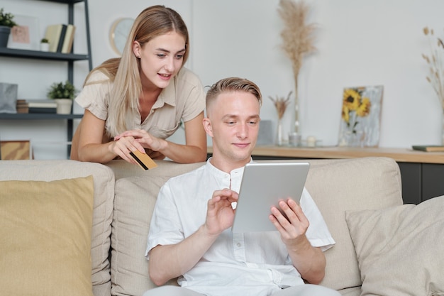 Jonge vrouw met creditcard leunend op de bank waar haar man met touchpad zit en door goederen in de online winkel bladert