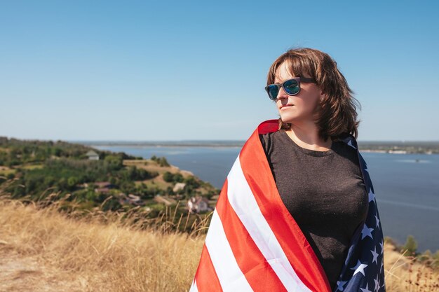 Jonge vrouw met de vlag van Amerika en met een zonnebril op de natuur op een zonnige dag Vrije levensstijl