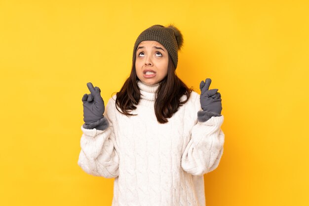 Jonge vrouw met de winterhoed over geïsoleerde gele achtergrond die met vingers kruisen en het beste wensen