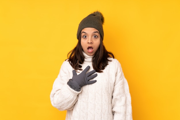 Jonge vrouw met de winterhoed over gele muur die verrast en wordt geschokt terwijl het kijken net