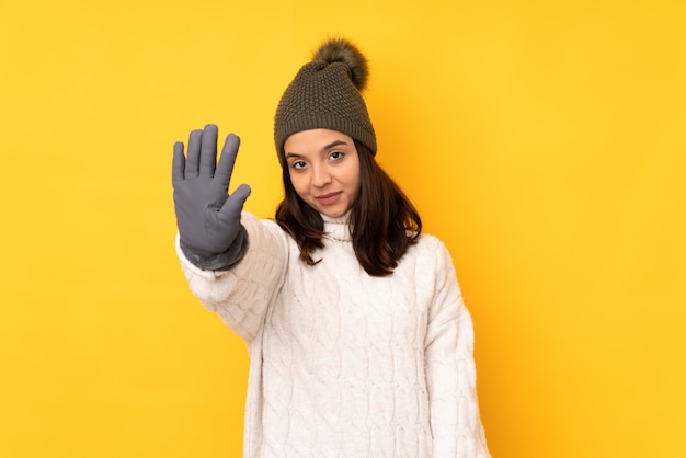 Jonge vrouw met de winterhoed over gele muur die vijf met vingers tellen