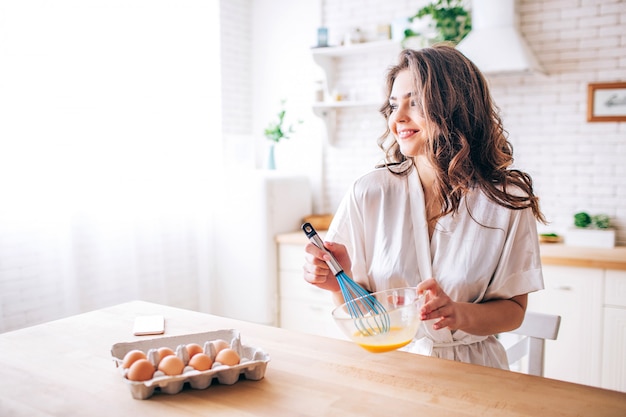 Jonge vrouw met donkere haartribune in keuken en het koken. Eieren mengen. Alleen. Ochtend daglicht. Kijk recht en glimlach. Telefoon op tafel.