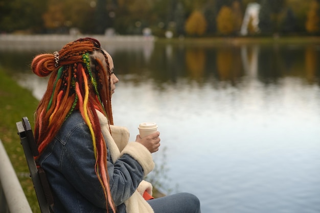 Jonge vrouw met dreadlocks in warme kleren zit in de buurt van meer koffie drinken in park