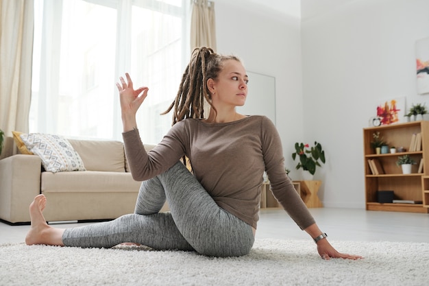 Jonge vrouw met dreadlocks zittend op de vloer in een van yoga-posities tijdens het strekken van arm- en beenspieren