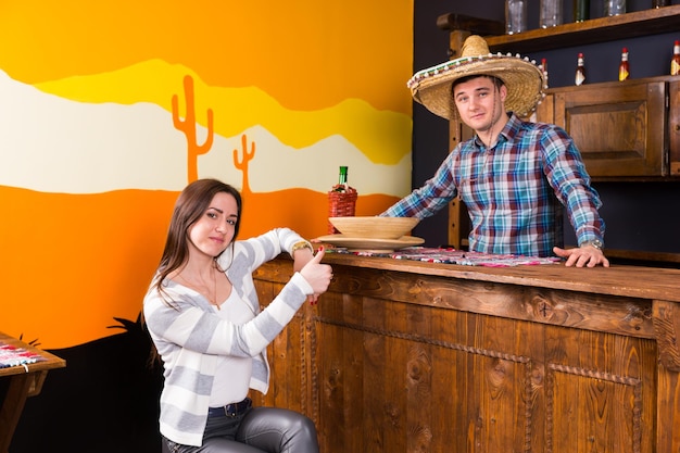 Jonge vrouw met duimen omhoog zittend aan de toog naast de barman met sombrero en een geruit hemd in een mexicaanse bar