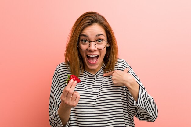 Jonge vrouw met een aardbei verrast wijzend op zichzelf, breed glimlachend.