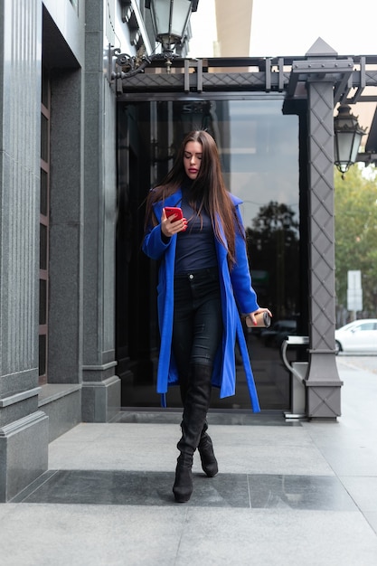 Jonge vrouw met een blauwe jas drinkt koffie om te gaan en praat op straat aan de telefoon. Vergrendeld realtime medium shot