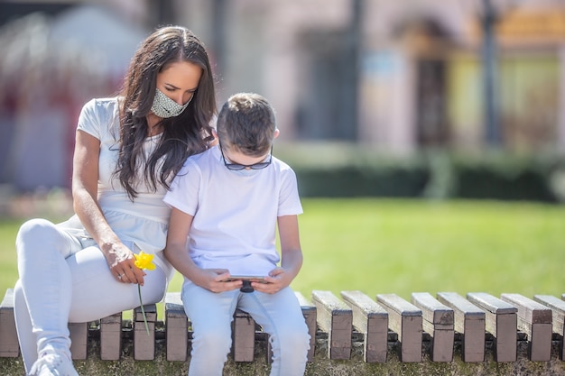 Jonge vrouw met een bloem zit naast een jongen in de stad, beide kijkend in een mobiele telefoon in zijn handen en gezichtsmaskers dragend.