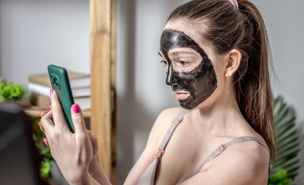 Jonge vrouw met een cosmetisch zwart masker op haar gezicht gebruikt een mobiele telefoon
