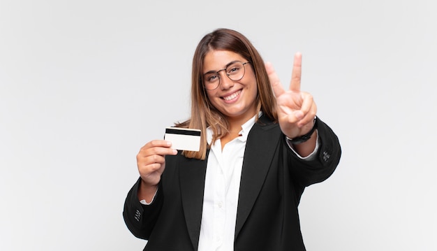Jonge vrouw met een creditcard die gelukkig, zorgeloos en positief glimlacht en kijkt, overwinning of vrede met één hand gebaart
