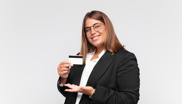 jonge vrouw met een creditcard die vrolijk glimlacht, zich gelukkig voelt en een concept in exemplaarruimte met handpalm toont