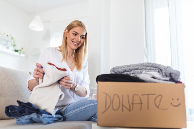 Jonge vrouw met een donatiedoos thuis Donatiedoos voor armen met kleding in vrouwelijke handen Vrouw geeft kleding aan een schuilplaats Vreugdevolle vrouw houdt een doos met gedoneerde kleding vast