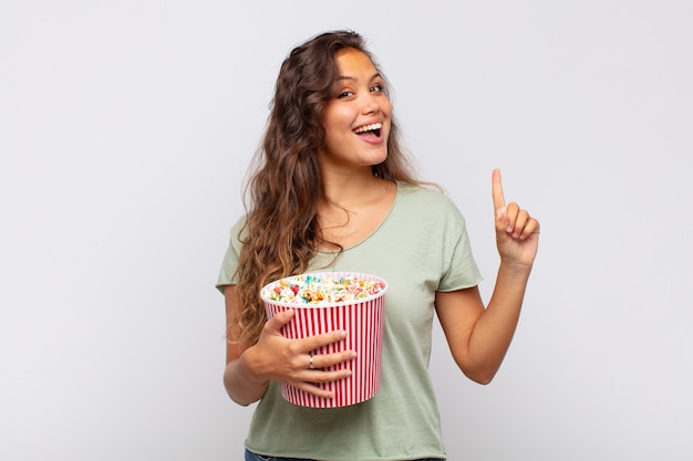 Jonge vrouw met een emmer met popconrs die zich als een gelukkig en opgewonden genie voelt na het realiseren van een idee, opgewekt de vinger opstekend, eureka!