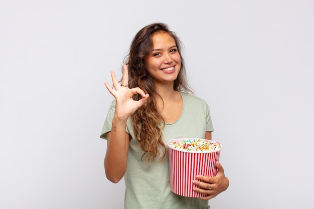 Jonge vrouw met een emmer met popconrs die zich gelukkig, ontspannen en tevreden voelt, goedkeuring toont met een goed gebaar, glimlachend