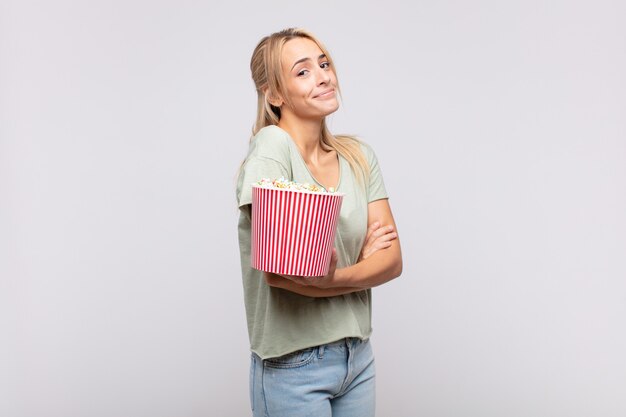 Jonge vrouw met een emmer met popcorn die haar schouders ophaalt, zich verward en onzeker voelt, twijfelt met gekruiste armen en verbaasde blik