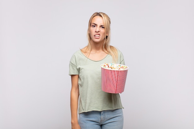 Jonge vrouw met een emmer met popcorn die zich verbaasd en verward voelt, met een domme, verbijsterde uitdrukking op zoek naar iets onverwachts