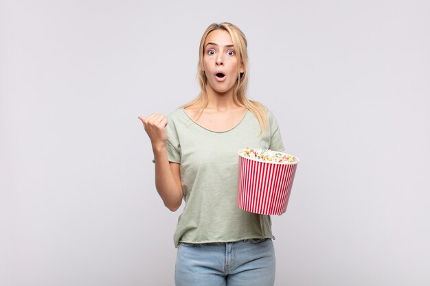 jonge vrouw met een emmer met popcorn kijkt verbaasd van ongeloof, wijst naar een voorwerp op de zijkant en zegt wow, ongelooflijk