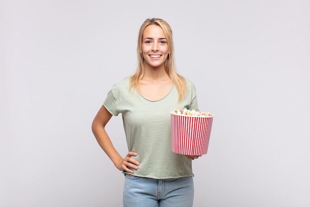 Jonge vrouw met een emmer popcorns die gelukkig met een hand op heup en zelfverzekerde, positieve, trotse en vriendelijke houding glimlacht