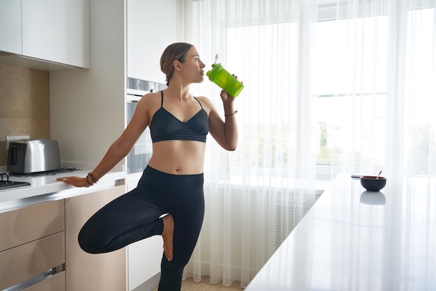 Jonge vrouw met een fles eiwitshake aan tafel in de keuken na thuistraining