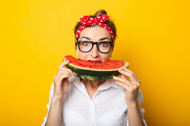 Jonge vrouw met een glimlach in een rode hoofdband en glazen eet een watermeloen op een gele muur.