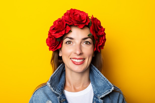 Jonge vrouw met een glimlach in een spijkerjasje en een krans van rode bloemen op haar hoofd op een gele muur.