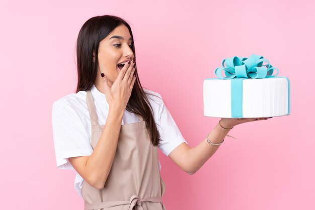 Jonge vrouw met een grote cake over geïsoleerde muur met verrassing en geschokte gelaatsuitdrukking