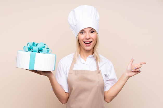 Jonge vrouw met een grote cake over geïsoleerde muur verrast en wijzende vinger aan de kant