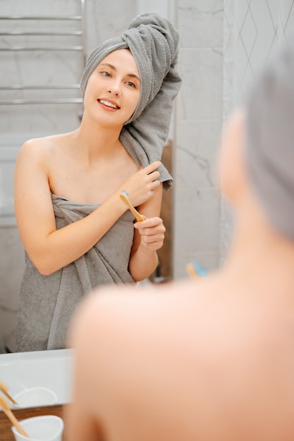 Jonge vrouw met een handdoek op haar hoofd poetst haar tanden met een eco-borstel in de badkamer. Concept van tandheelkunde en zorg.