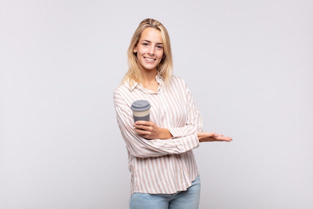 Jonge vrouw met een koffie die vrolijk glimlacht, zich gelukkig voelt en een concept in exemplaarruimte met handpalm toont