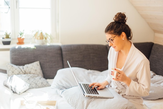 Jonge vrouw met een koffiekop met een laptop terwijl ze thuis op bed zit