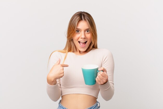 Jonge vrouw met een koffiemok die zich gelukkig voelt en naar zichzelf wijst met een opgewonden