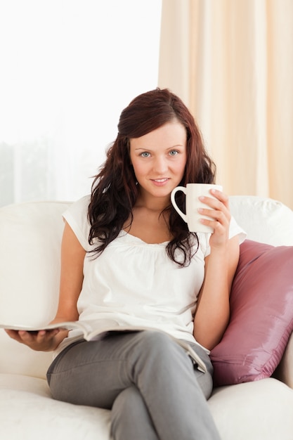 Jonge vrouw met een kop en een tijdschrift op zoek naar de camera