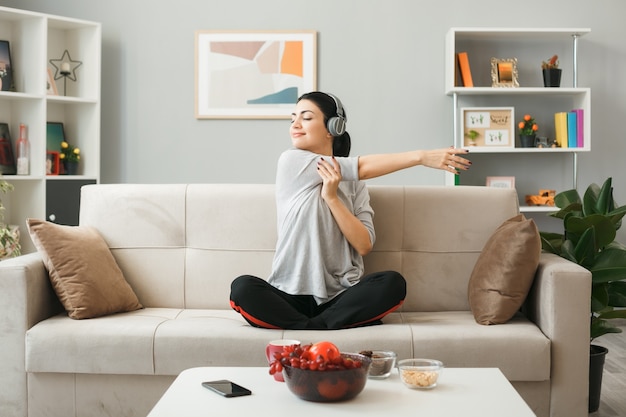 Jonge vrouw met een koptelefoon die yoga doet, zittend op de bank achter de salontafel in de woonkamer