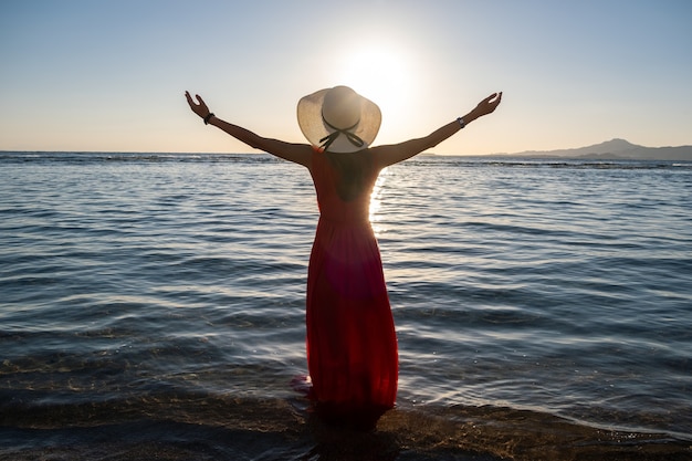 Jonge vrouw met een lange rode jurk en een strohoed die de handen in het zeewater op het strand opsteekt en geniet van het uitzicht op de rijzende zon in de vroege zomerochtend.