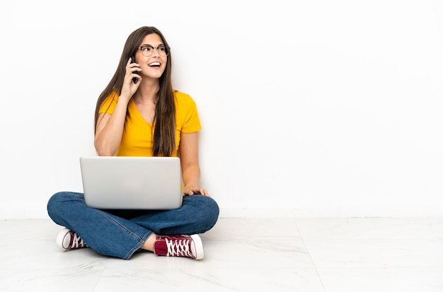 Jonge vrouw met een laptop die op de grond zit en een gesprek voert met de mobiele telefoon