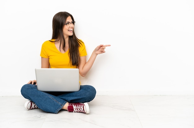 Jonge vrouw met een laptop die op de grond zit en met de vinger naar de zijkant wijst en een product presenteert