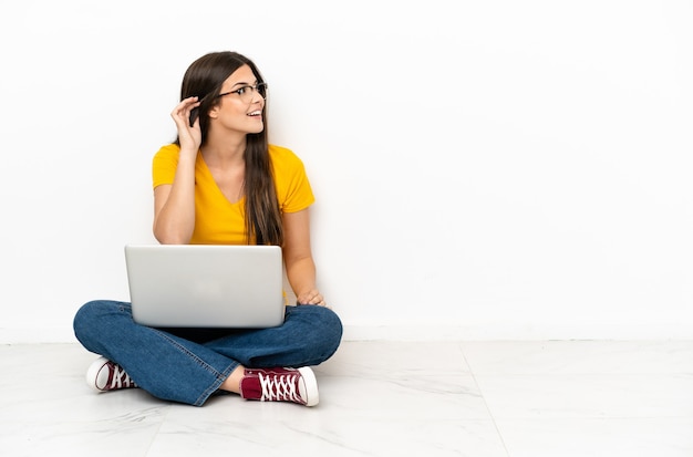 Jonge vrouw met een laptop die op de grond zit te luisteren naar iets door de hand op het oor te leggen