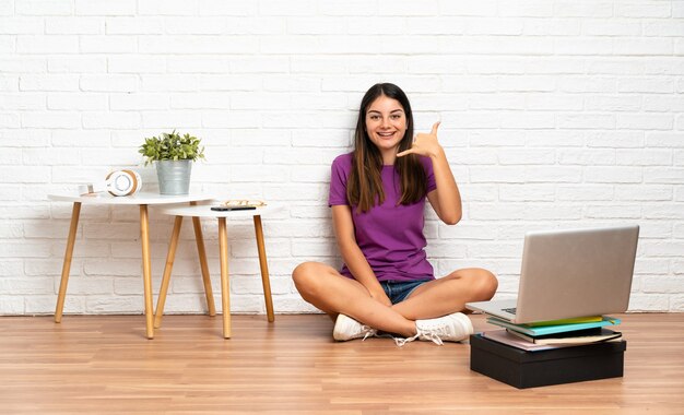 Jonge vrouw met een laptop zittend op de vloer bij binnenshuis telefoongebaar maken. Bel me terug teken