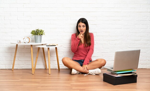 Jonge vrouw met een laptop zittend op de vloer binnenshuis verrast en wijzend naar voren